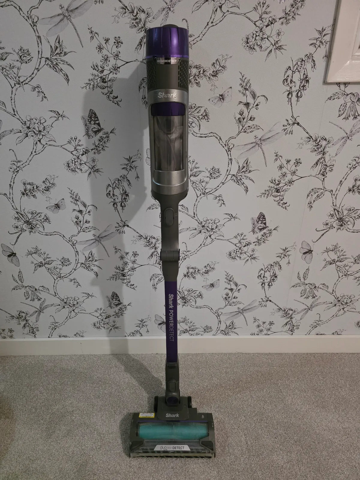 Shark PowerDetect cordless stick vacuum standing on grey carpet against floral wallpaper